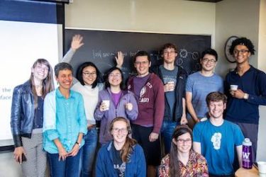 A large group of people pose in front of a black board. One person is raising her arms in celebration.