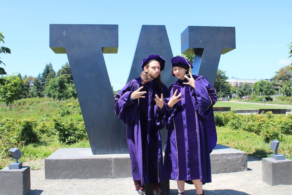 Ashley Ruba and James Clark stand in front of metal W sign outdoors in purple graduation robes