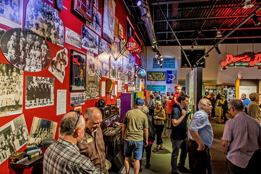 People gather inside museum. Historical posters adorn wall.